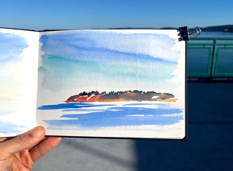 a hand holding a sketchbook with a watercolor sketch of an island with sky above and loosely painted water below