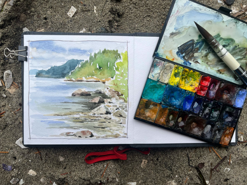 A sketchbook on the sand with a watercolor palette and paint brush overlaying it.