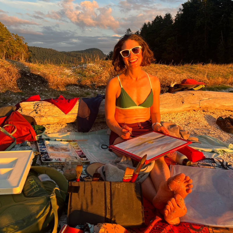 A woman sketching on the beach with watercolor supplies around her.