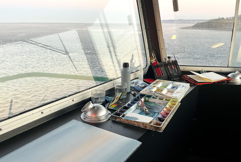Art supplies laid out on a countertop in front of large windows overlooking the sea