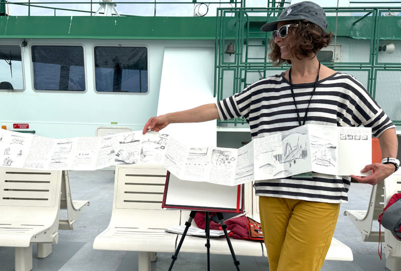 A woman wearing a hat in a striped shirt and yellow pants holding a sketchbook