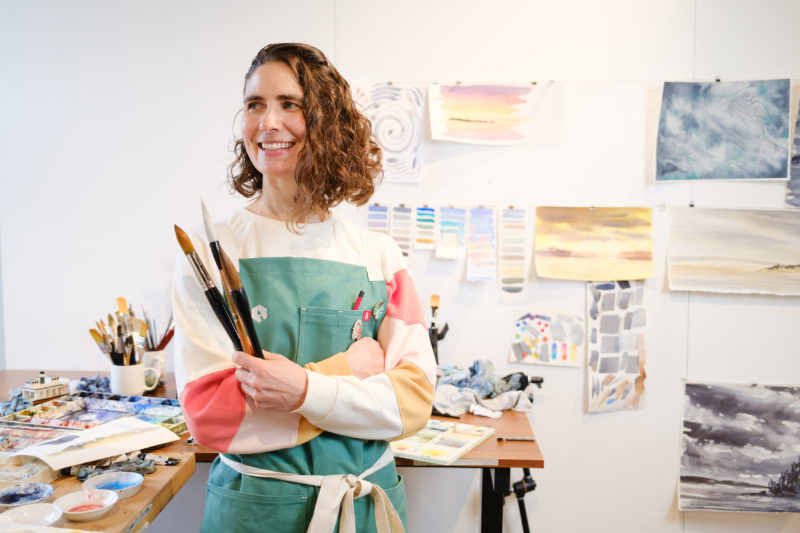 A photo of a woman in a green apron and brown curly hair smiling and holding large paint brushes.